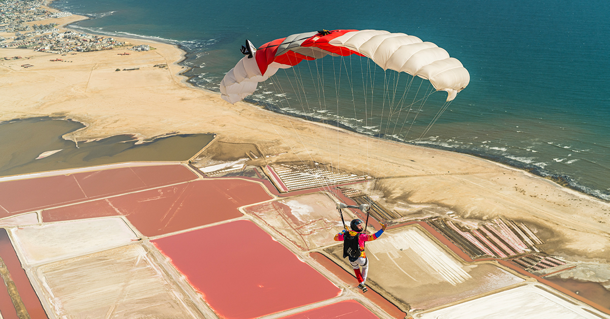 Skydiving coast under an open chute above the read fields and pale sands of the Namibia coast.